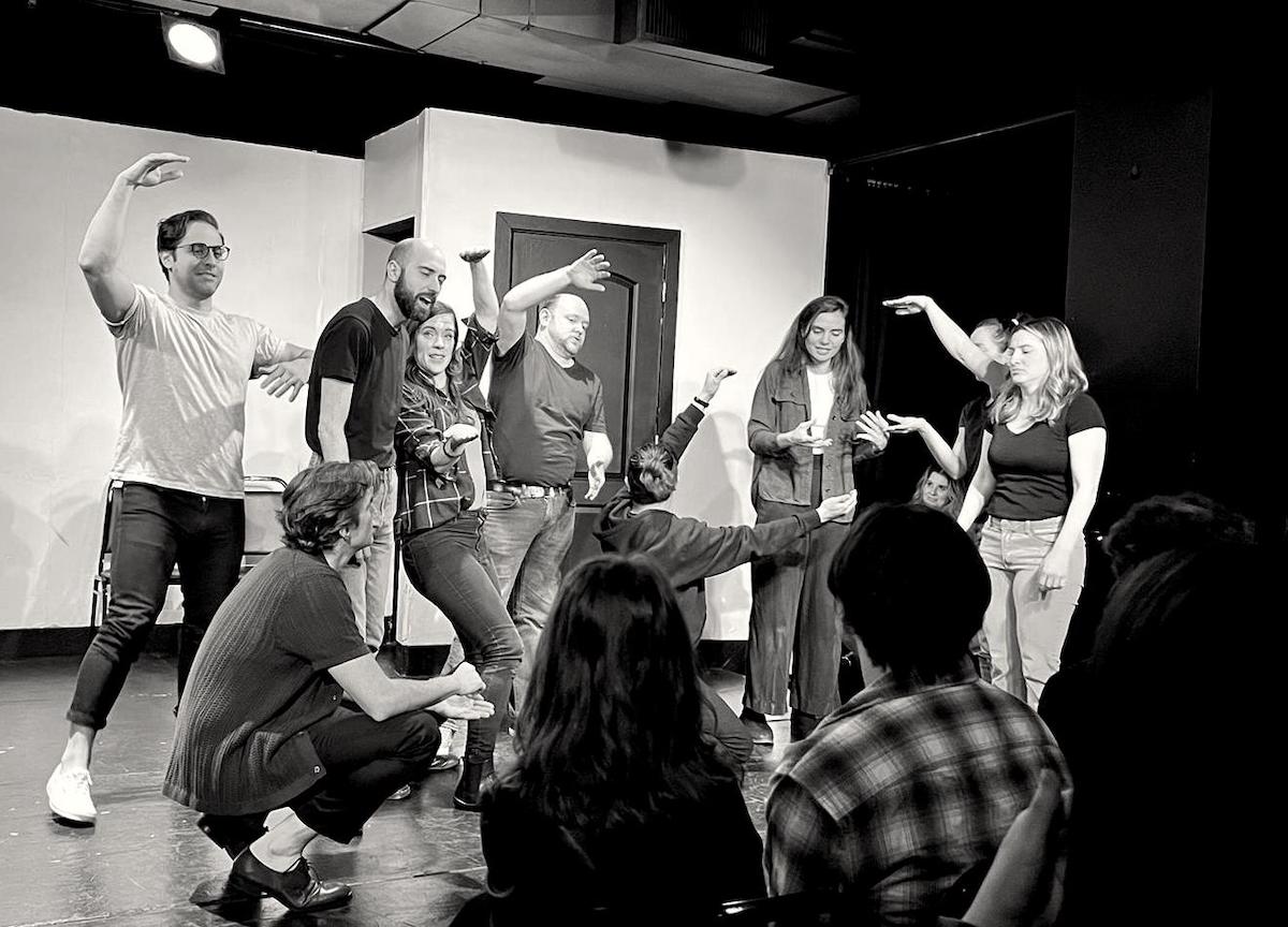 Black and white photo of 9 improvisers in various striking poses on a small black box theater stage